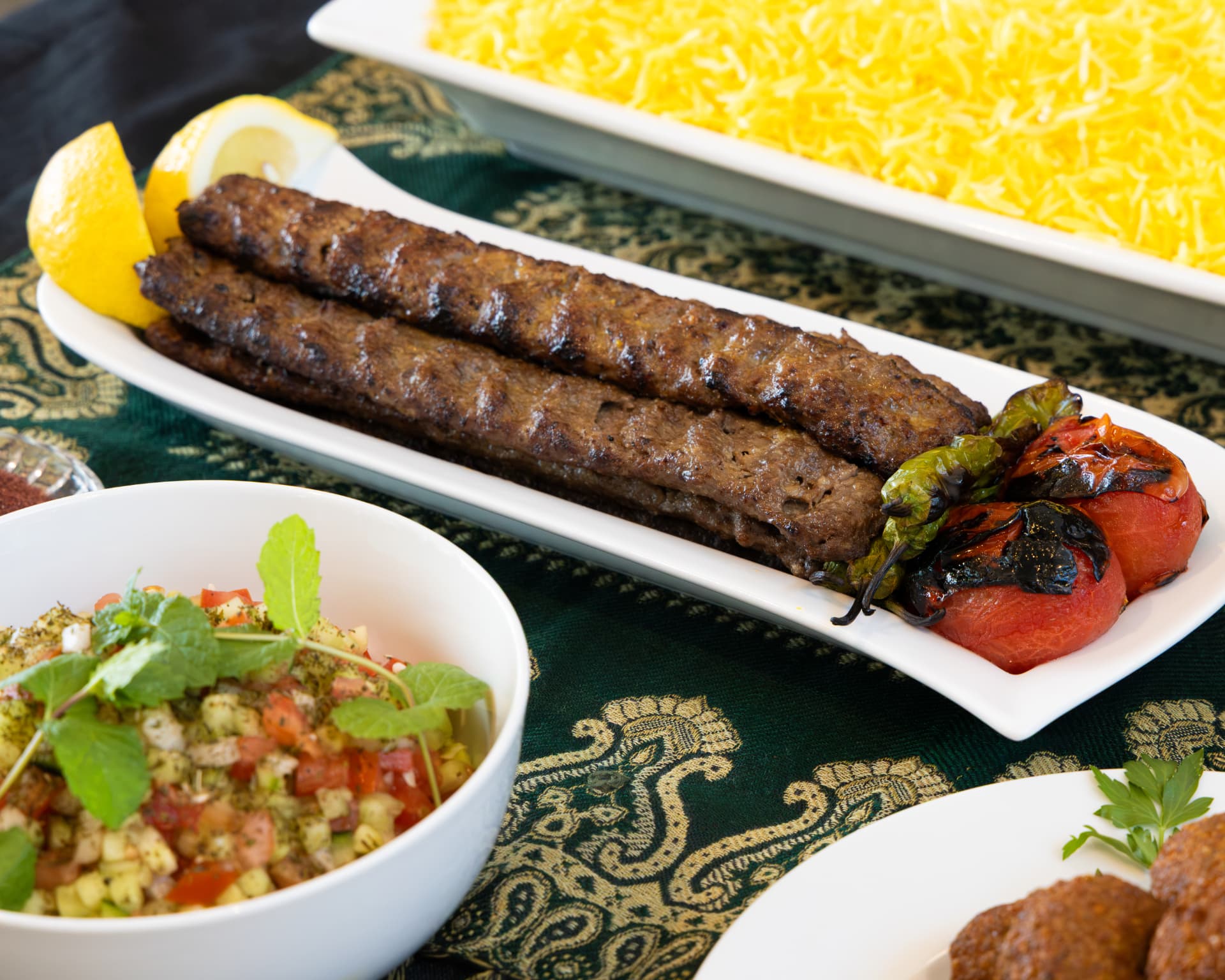 Beef koobideh kabob with saffron rice, grilled tomatoes, and salad Shirazi on a set table