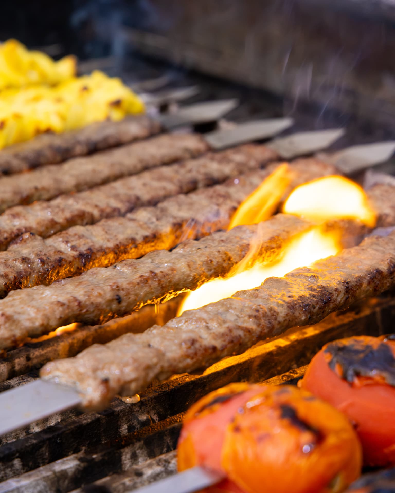 Koobideh kabobs on flat skewers over open flame, with joojeh and grilled tomatoes